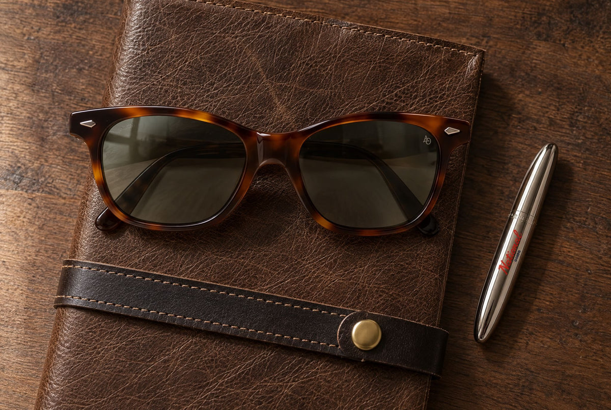 American Optical Saratoga sunglasses in tortoise resting on a Local Branch leather journal beside a National Goods x Fisher bullet pen, seen from above